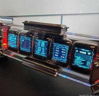 Front view of the Times Gate device displaying multiple screens including time, stock market data, and audio levels, showing blue-themed pixel art graphics.