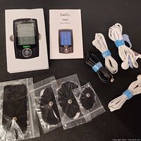 Photo of the Belifu TENS EMS device in foam packaging with instruction manual and multiple cables and electrode pads laid out flat on black surface.