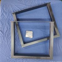 Two modern steel furniture legs with rectangular frame design and black satin finish lying flat on blue fabric, with pack of mounting hardware in the center.