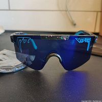 Front view of Pit Viper polarized sunglasses showing blue reflective lens with Pit Viper logo, including extra ear hooks in background.