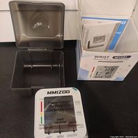 White MMIZOO wrist blood pressure monitor with display showing protective film and interface buttons. Black protective carrying case behind and original box with manual on side.