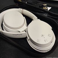 White Srhythm foldable over-ear headphones in black zippered case with control buttons visible on right ear cup.