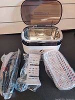 Ultrasonic jewelry cleaner with accessories including power cord, plastic basket, and inserts in packaging, placed in front of the unit with lid open.