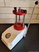 Full view of the waterproof pressure tester machine with white base, transparent cylindrical chamber with red caps, pressure gauge on the front, and lever on the side.