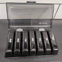 Open view of 7 black medication organizer cases, each marked with a day of the week, showing compartments for day and night medications.
