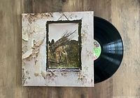 Photo showing the front cover of the Led Zeppelin untitled LP with a man carrying sticks in a framed picture on wallpaper background; vinyl record partially pulled out.