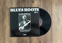 Vinyl LP record partially pulled out of the album sleeve. The album cover is black with white text 'BLUES ROOTS VOL. 8' and a black and white photo of Lonnie Johnson playing guitar. The vinyl label is visible with the Nig's Burlap Records logo and text.