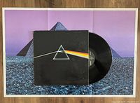 Photo showing the Dark Side Of The Moon vinyl LP partially out of its album jacket, which features the iconic prism and rainbow design, placed on top of a large 6-panel poster with blue and purple pyramid artwork.