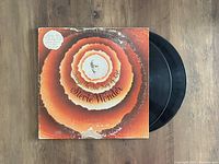 Photo showing front of album cover with circular sunburst design, Stevie Wonder name and title, along with two vinyl discs partially outside the cover on wooden surface.