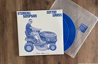 Photo showing the front cover of the gatefold vinyl record sleeve with a man riding a lawn mower in blue illustration, alongside two translucent blue vinyl discs partially visible and a clear plastic protective sleeve with a blue sticker.