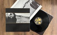 U2 The Joshua Tree vinyl LP record partially out of sleeve, visible black vinyl disc with yellow label, original album cover showing band photo in desert landscape, and black and white liner notes insert.