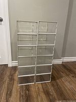 Stack of 12 clear plastic vented shoe display boxes stacked vertically in two columns of six each on a wooden floor against a wall. Each box has a front door with ventilation holes and patterned surface.