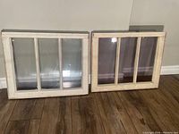 Two antique three-paned wooden windows with white painted frames showing chipping, placed upright against a wall on a wooden floor