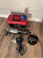 Red and black toolbox with assorted tools including oil can, metal disc sander, and electric drill with cord