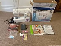 Photo showing Brother JX2217 sewing machine, box, and assorted accessories laid out including a sewing kit, thread, needles, and instructional book.