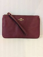 Front view of the burgundy leather wristlet showing textured leather, gold Coach logo, and attached wrist strap.