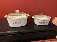 Two Corning Ware Cornflower Blue casserole dishes with glass lids, one 1.5 litre and one 12 cup, displayed side by side on a black surface against a red wall.