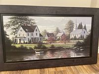 Framed print showing serene landscape with houses beside water, framed in dark wooden frame with some wear.