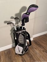 Set of six junior left handed golf clubs in a black and white Power Built golf bag standing upright against a wall on wooden floor.
