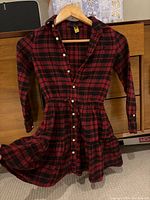Photo of the red and black plaid button-up girls dress with long sleeves on a wooden hanger, displayed in indoor setting.