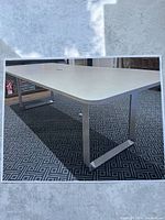 Photo showing modern office desk with dove grey tabletop and chrome legs on patterned carpet. Light and shadow indicate smooth surface and metal legs. Cable management cutout visible in center.