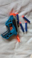 Blue and gray Nerf blaster with orange trigger next to 4 blue foam darts with orange suction cup tips on a fabric background.