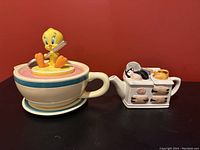 Photo showing both the Warner Brothers Studio Store Tweety Bird teapot with matching plate and the cat themed tea pot by Giftcraft side by side on black surface with red background.