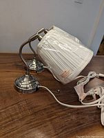 Two IKEA plug-in wall sconces with brushed nickel curved curved metal bases and cream pleated fabric shades, one shade unopened in plastic, one with minor sun discoloration. White cords with pull chains and plugs are visible, laid on wood surface.