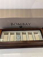 Closed rectangular solid wood watch display case with beveled glass top showing interior watch cushions and Bombay logo on glass.