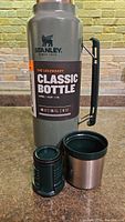 Side view of the Stanley Classic Bottle thermos with stopper and cup lid placed on counter, label showing capacity and insulation times.