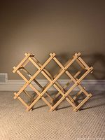 Photo of expandable foldable wooden wine rack on carpet, light wood color, empty, showing diamond grid structure for 10 bottles.