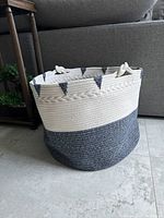 Photo showing the textile woven basket placed on a tiled floor in front of a grey couch, highlighting its blue and white woven pattern and handle on the rim.