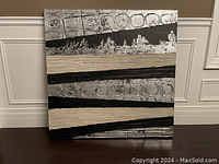 Front view of square textured abstract wall art with beige, black, and silver tones, showing horizontal stripes and circular pattern detail.