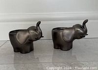 Pair of pewter-colored elephant candle holders on tiled floor, showing side profile with trunks raised.