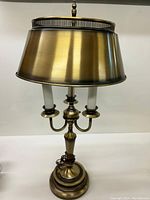 Frontal photo showing the gold Florentine metal tole drum shade, three faux candle arms, and base with a cord coiled at the bottom.