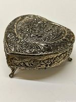 Closed heart-shaped vintage silver-colored jewelry box showing embossed floral and rose pattern on lid and sides with curved feet.
