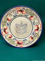 Full view of ceramic plate with pomegranate and leaf motif on rim and coat of arms in center, with blue trim on edge.