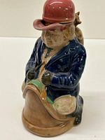Front view of the ceramic decanter shaped as a seated man with a red hat and blue coat, holding a pipe, with a cat on his back.