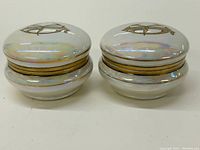 Pair of small round ceramic jars with lids side by side showing iridescent white finish and gold character on top