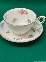 Teacup resting on saucer showing side profile of cup with floral pattern and gold edging on green background.