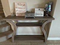 Console table against a wall with decorative items on top including a sign, metal box, glass jar with potpourri, and floral canvas art.