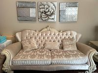 Full frontal view of beige French style sofa with button tufted back and 3 decorative cushions under 3 mounted canvas wall art pieces.