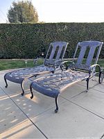 Pair of iron chaise lounges made of dark metal with lattice seats, backs, and decorative scroll arms placed on outdoor tiles with bushes in background