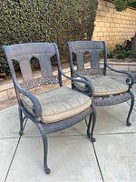 Pair of dark iron outdoor chairs with detailed backrest design and beige cushions placed on a patio with brick wall and greenery background