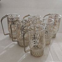 Six clear cylindrical glass mugs featuring a gold flake mottled pattern with gold-tone metal handles and bases, arranged on a flat surface.