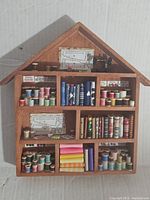 Front view of vintage sewing themed shadow box shaped like a small house filled with colorful wooden spools of thread, folded fabric, and sewing tools.