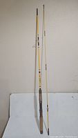 Full view of two-piece yellow vintage Sportfisher fishing rod with cork handle, metal reel seat, and line guides placed upright on a white surface against a wall.