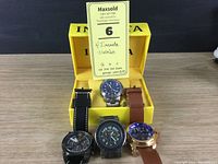 Four Invicta watches displayed in an open yellow Invicta-branded box on a wooden surface. Two watches have leather straps (one black, one brown), two have stainless steel link bracelets.
