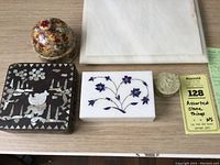 Photo showing five assorted stone and wood decorative items on a wooden surface including a round domed stone box with floral design, a white stone box with blue floral inlay, a small carved stone scent holder, a black wood and lacquer box with mother of pearl insets, and a white marble rectangular plinth.