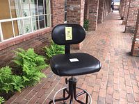 Full view of black drafting stool showing padded seat, backrest, and base with five wheels.